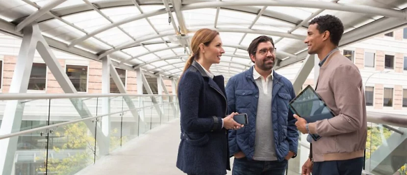 Three person are standing and one of them is holding tablet is his hand and they all are discussing